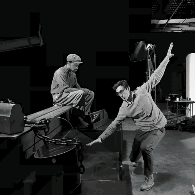 Two men in a recording studio, one sitting on a piano, the other leaning forward with one arm raised and the other hand on the piano, both wearing glasses and casual clothing.