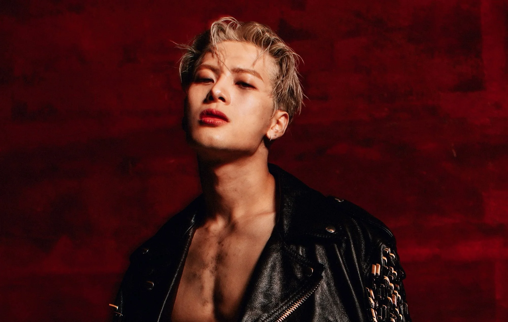 Person with short blonde hair, wearing a black leather jacket, posing against a dark red background.