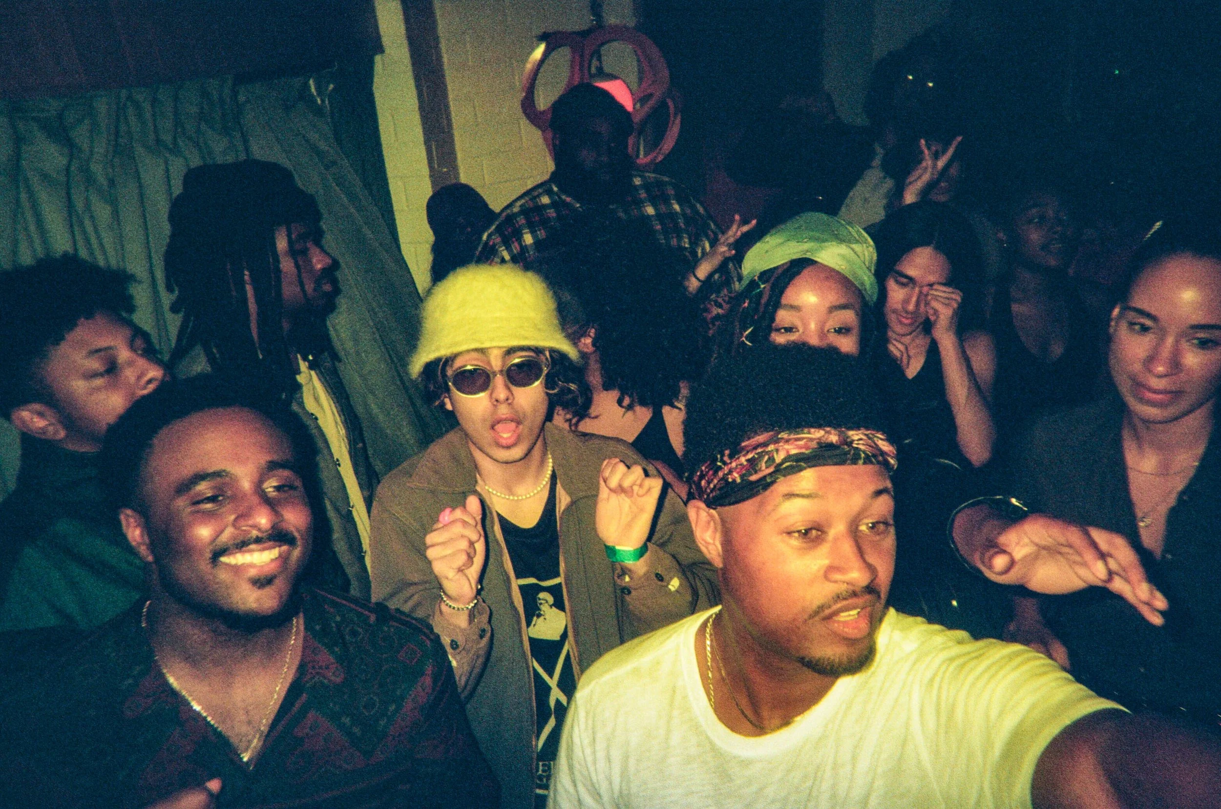 A group of young adults at a party or gathering, smiling and taking a selfie. They are dressed casually, with some wearing sunglasses, hats, and accessories, and one person making a peace sign.