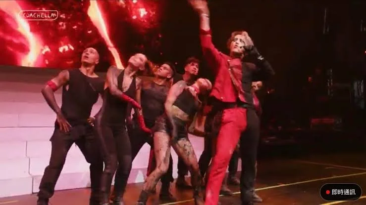 Group of six performers on stage, with one singer in red outfit leading the performance, during a concert with a fiery background.