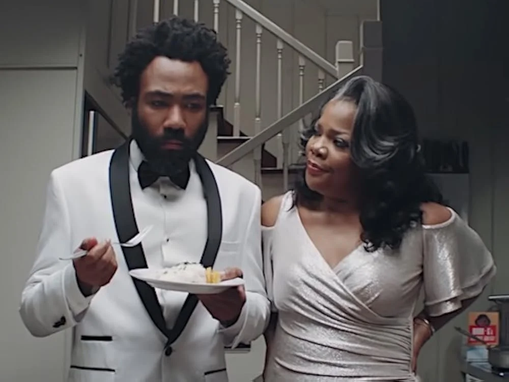 A man in a tuxedo holding a plate of food stands next to a woman in a light-colored dress, both inside a house near a staircase.