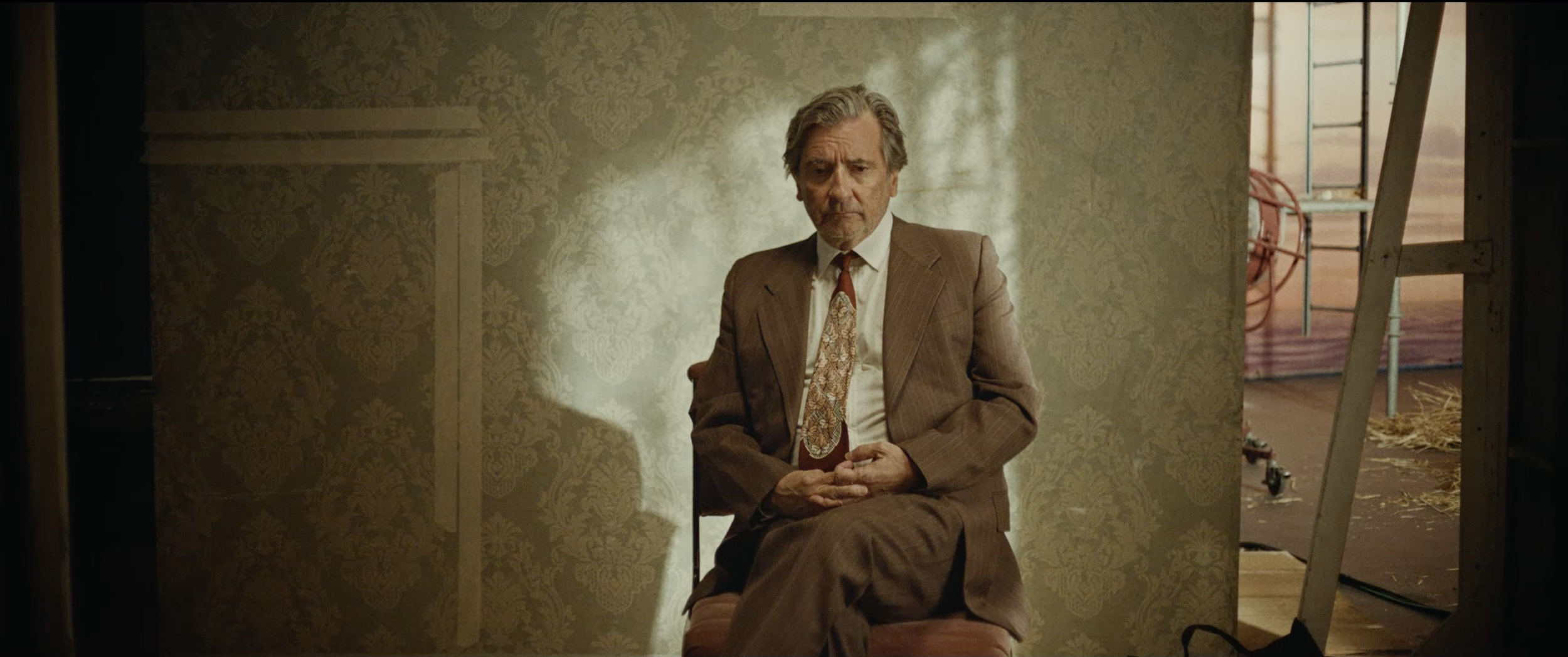 A man in a brown suit with a patterned tie sitting on a chair against a textured wallpapered, Imploding the Mirage by The Killers, a short film directed by Sing J Lee, starring Griffin Dunne, Kansas Bowling