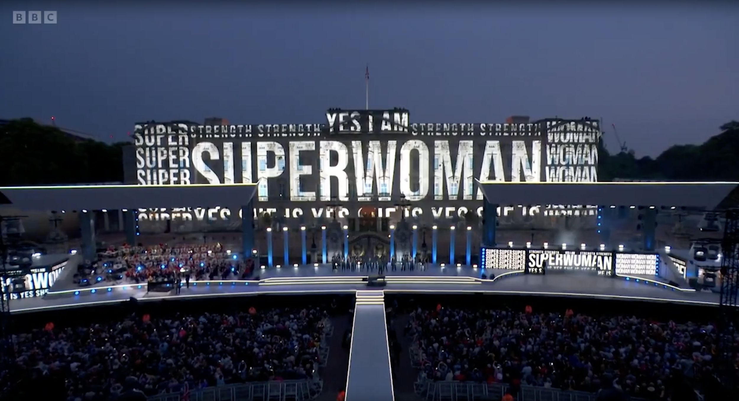 A large crowd gathered at a stadium or arena with a prominent sign displaying the words "SUPERWOMAN" and related phrases like "YES I AM" and "STRENGTH" on big screens. The event appears to celebrate or support women, with the sign and crowd indicating a rally or gathering focused on women's empowerment.