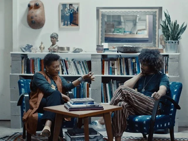 Two women having a conversation in a cozy room with bookshelves and decorative items, one woman speaking animatedly and the other listening thoughtfully.