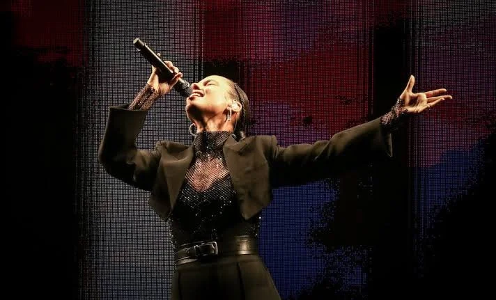 Performer singing on stage with microphone, arms outstretched, wearing a black blazer and glittery top, in front of LED backdrop.