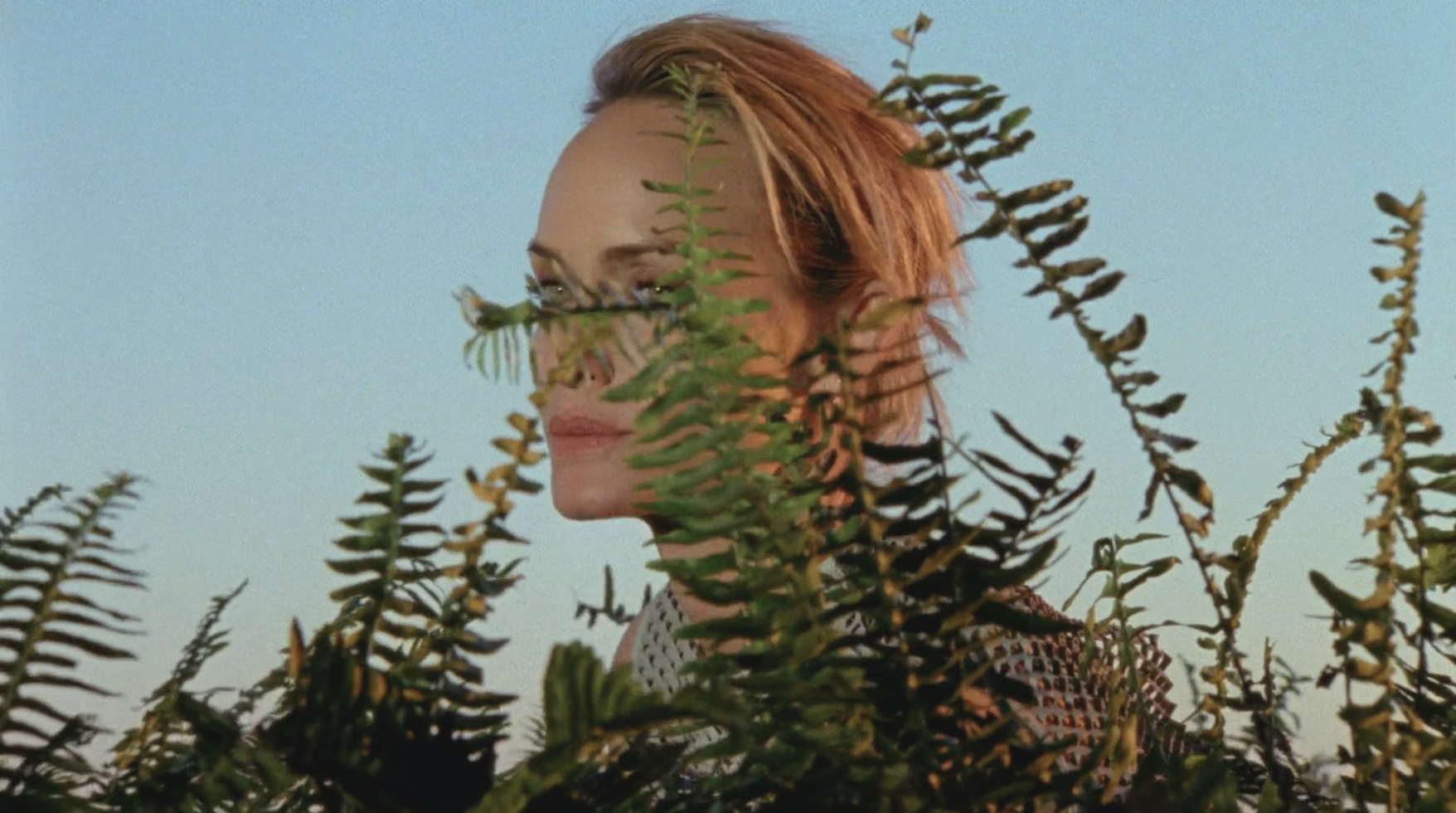 A woman with short red hair and makeup peeking through green fern plants against a clear sky.