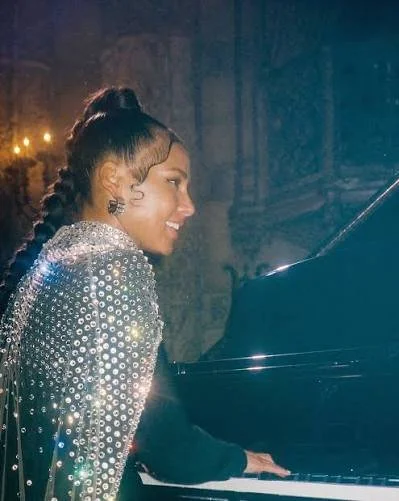 A woman with styled hair, wearing a sparkling, embellished outfit, playing a grand piano in a dimly lit, elegant setting.