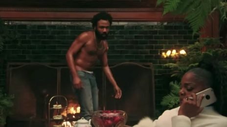 A shirtless man with dark hair and a beard is standing near a fireplace, while a woman with braided hair is sitting and talking on the phone in a room decorated with plants.
