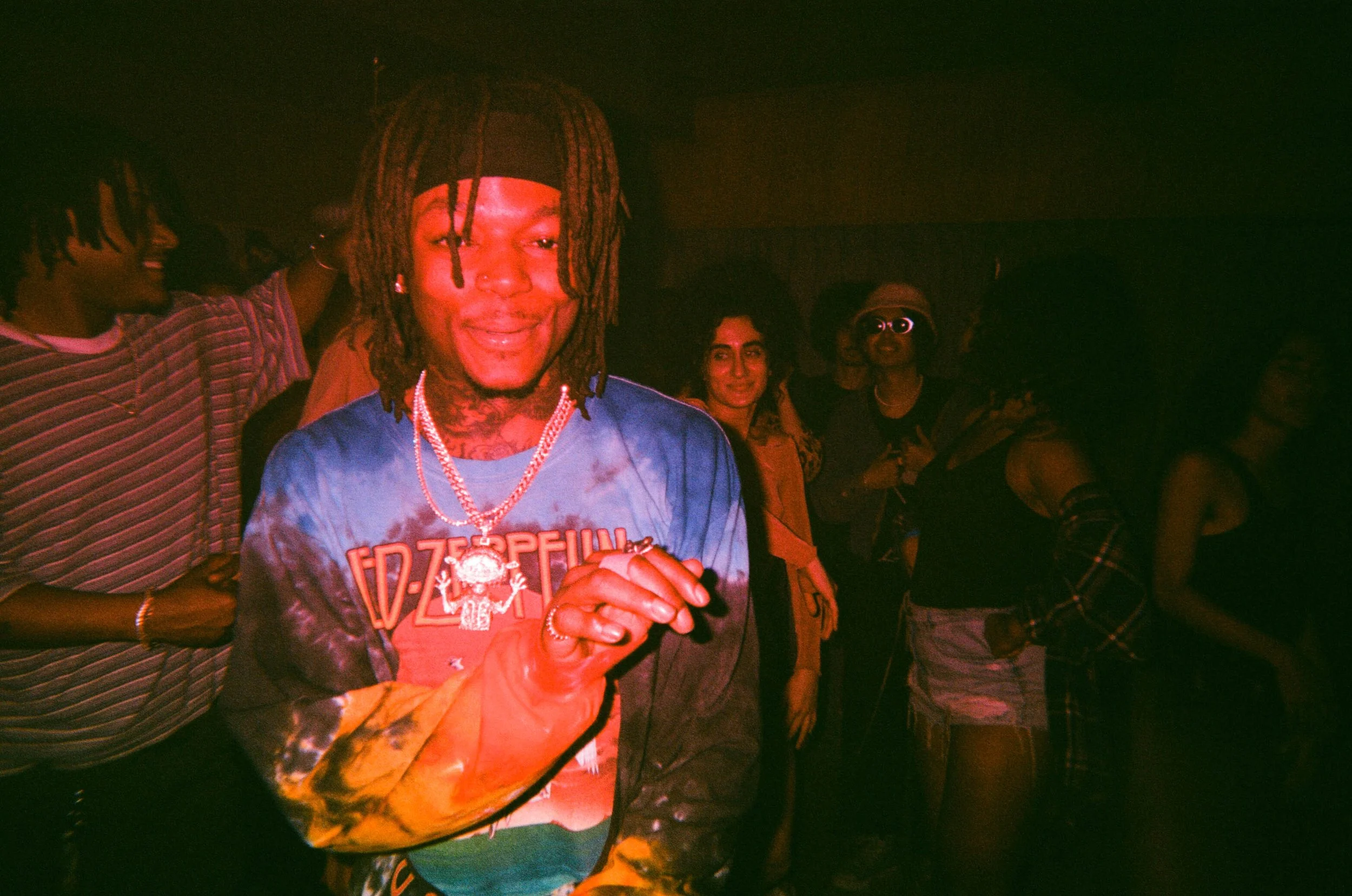 A group of people at a party or nightclub, with one person in the foreground wearing a tie-dye shirt and jewelry, smiling with eyes closed, surrounded by others in dim, colorful lighting.