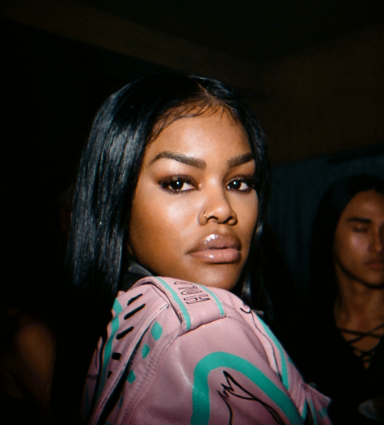 Close-up of a woman with long black hair, makeup, and a nose ring, wearing a pink and teal bag with writing on it, looking at the camera.
