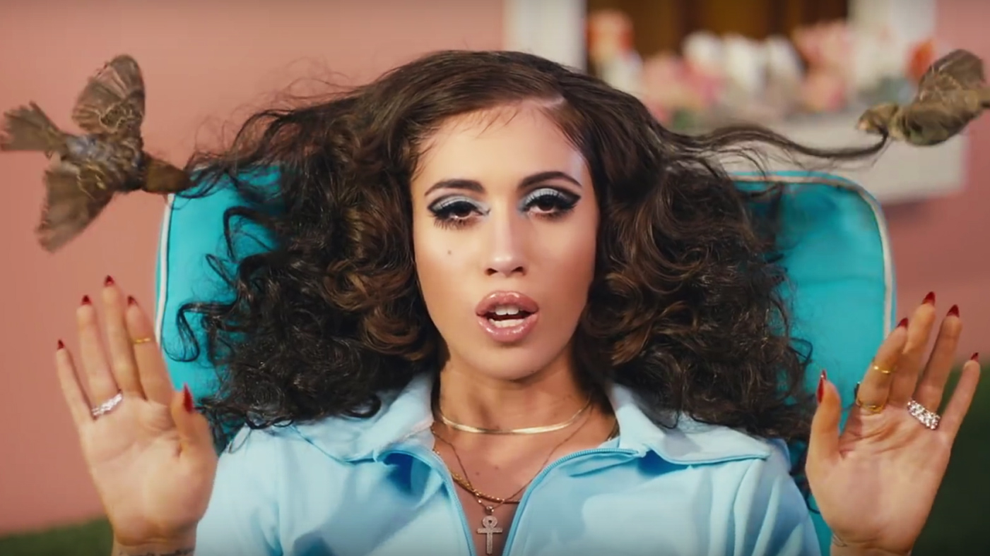 A woman with voluminous curly hair, wearing a light blue tracksuit, sitting in a turquoise chair with her hands raised, in front of a pink background with blurred objects. Kali Uchis After The Storm music video by Nadia Lee Cohen