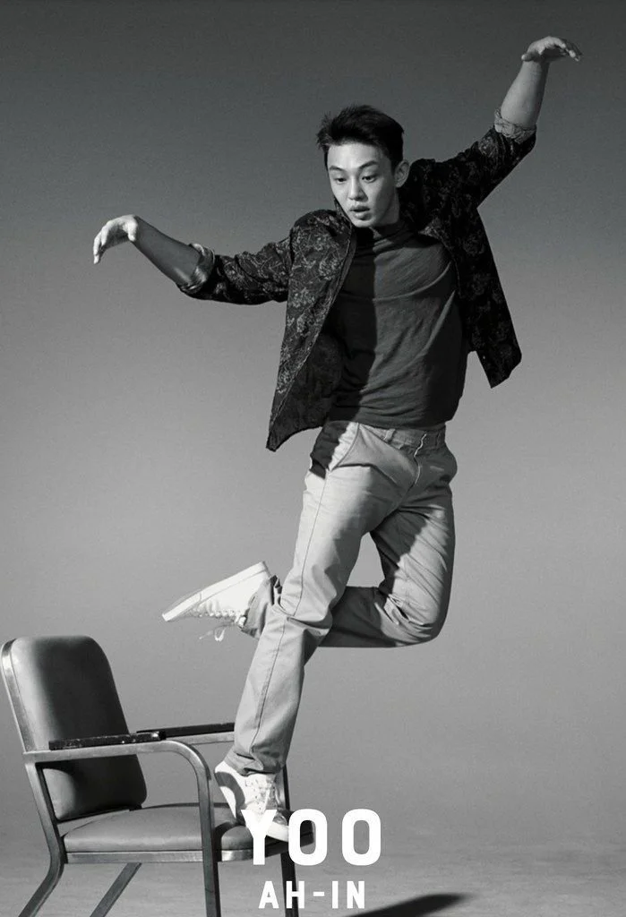 A young man is balancing on one leg on a chair while jumping, with arms extended. The photo is in black and white.
