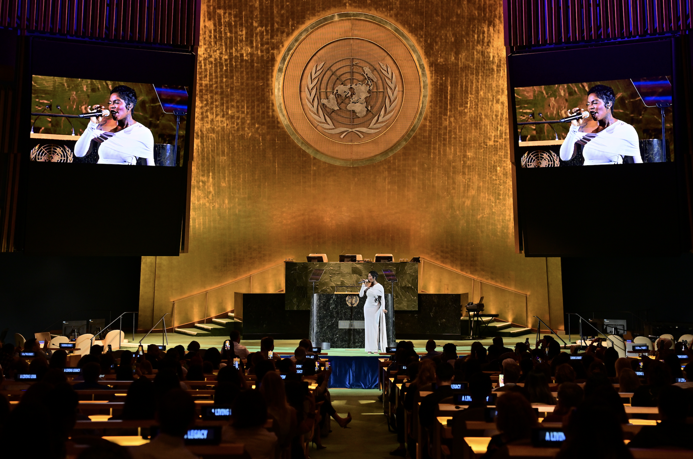 A woman in a white gown speaking at a podium inside the United Nations Assembly Hall, with large screens showing her image and the UN emblem behind her.