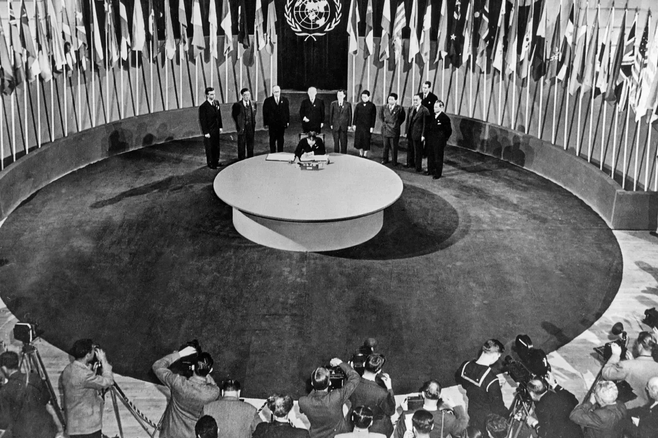 Historical black-and-white photograph of representatives signing the United Nations Charter at the UN Conference on International Organization, with photographers and journalists in the foreground.