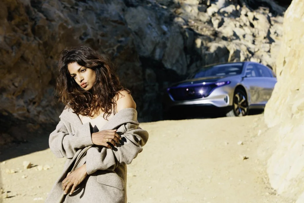 Woman with curly dark hair wearing a gray coat standing outdoors near rocky terrain with a silver car parked in the background.