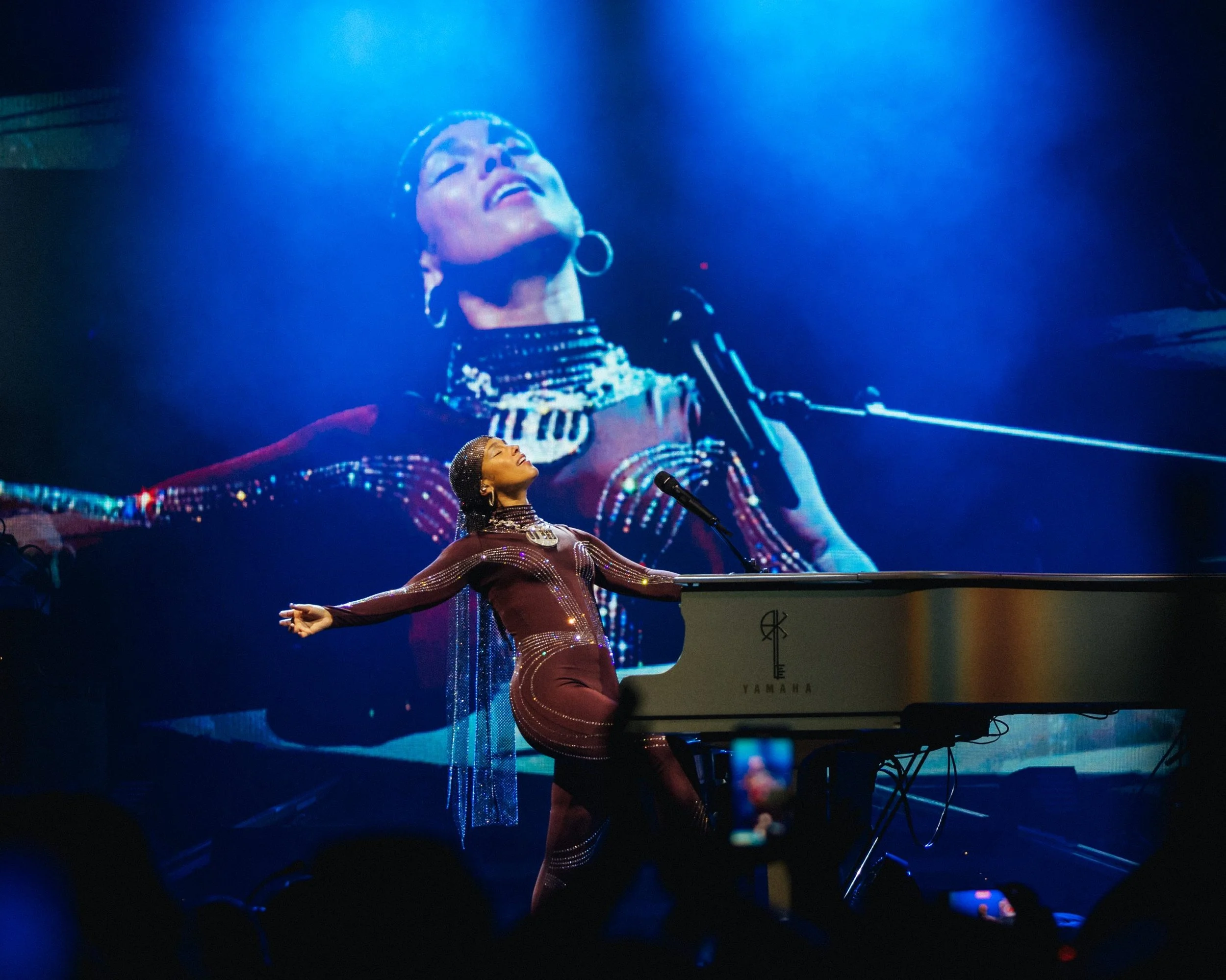A woman plays a Yamaha keyboard on stage, with a large screen behind her showing her image. The stage is lit with blue lighting, and the woman is wearing a sparkling outfit with arms outstretched. Alicia Keys World Tour for KEYS album.