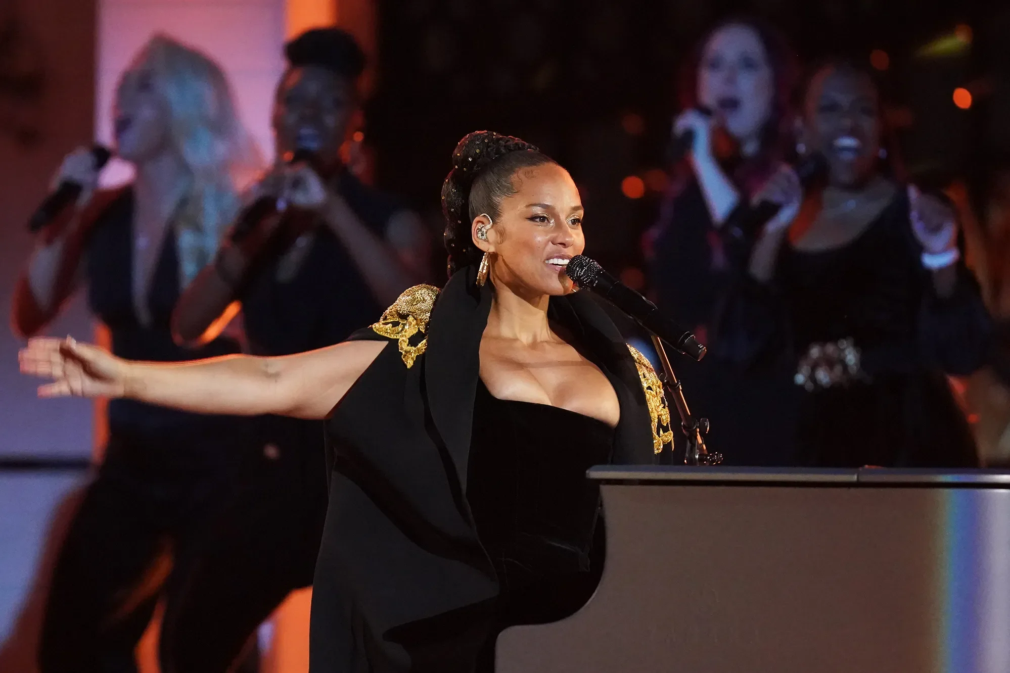 A woman performing at a concert, singing into a microphone with her arms outstretched, wearing a black dress with gold embellishments on the shoulders. In the background, a television screen displays other women singing.