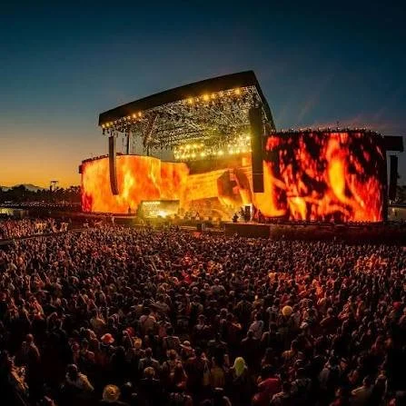 Large outdoor concert stage with bright lights and fiery screen displays at dusk, filled with a large audience.