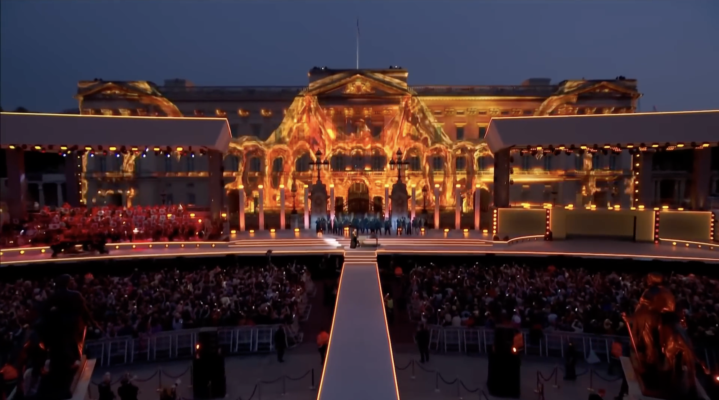 A large outdoor stage with bright, colorful lighting projections on a building in the background during a nighttime event. The stage has a crowd gathered in front of it.