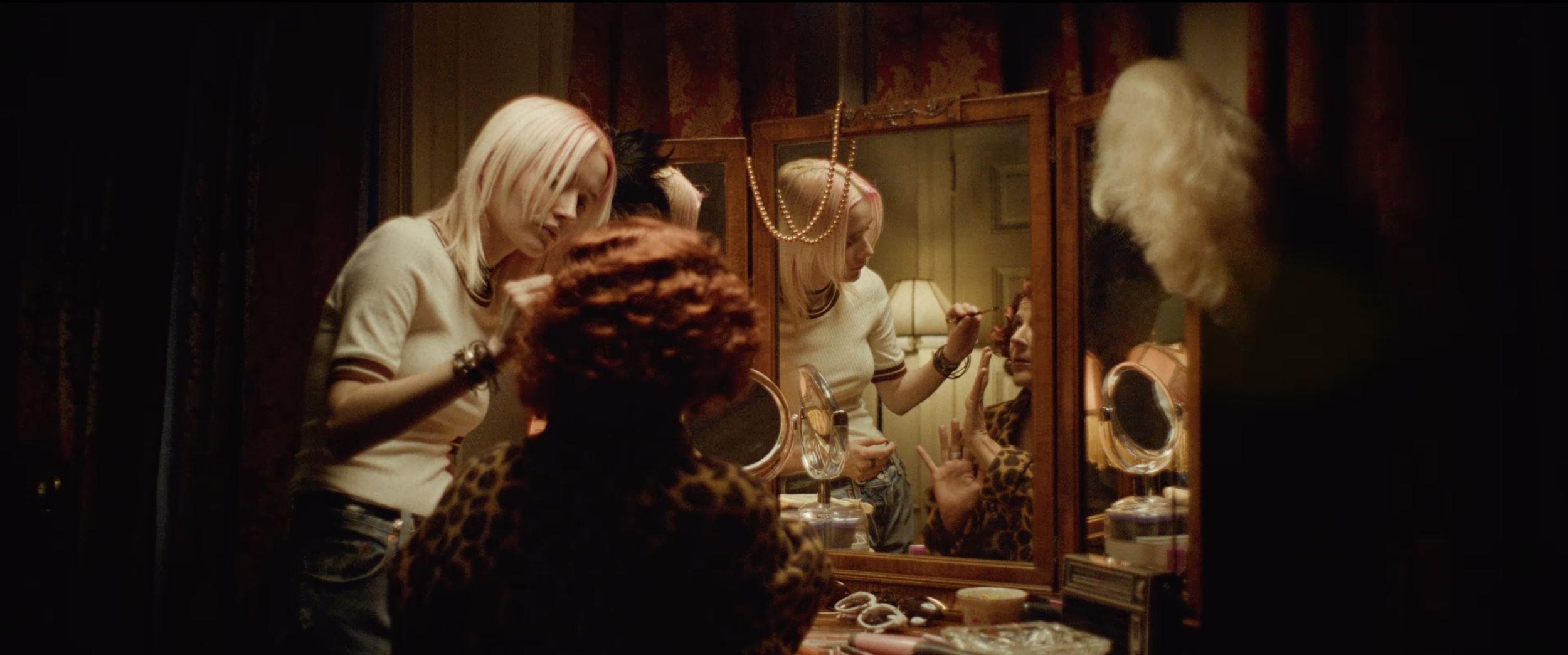 Three women in a dimly lit room are gathered around a mirror, applying makeup and styling hair. One woman with blonde hair is looking into the mirror while another with curly red hair, wearing a leopard print jacket, is also in front of the mirror. The third woman, with black hair, is partially visible. The vanity table holds various makeup and beauty products.
