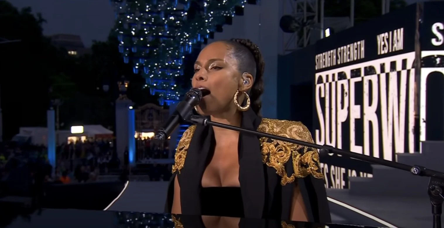 A woman singing into a microphone on a stage during an outdoor event at night. She is wearing large hoop earrings and a black and gold jacket with intricate detailing, with a sign in the background that says 'SuperW'.