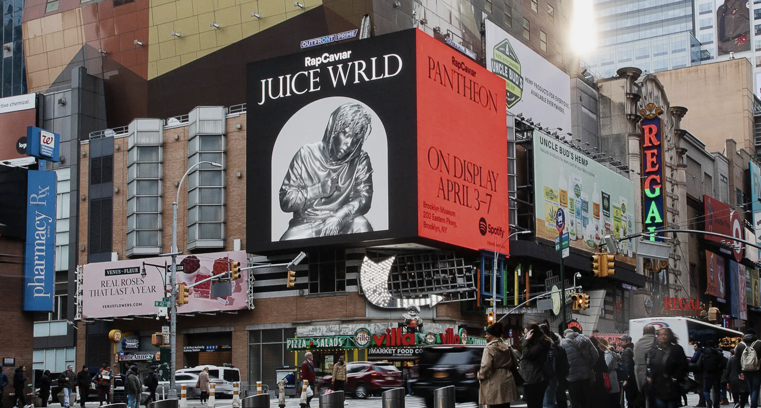City street scene with digital billboards, including a large one advertising Juice WRLD's art exhibit and a red one for RapCaviar. Pedestrians and vehicles are visible on the busy street.