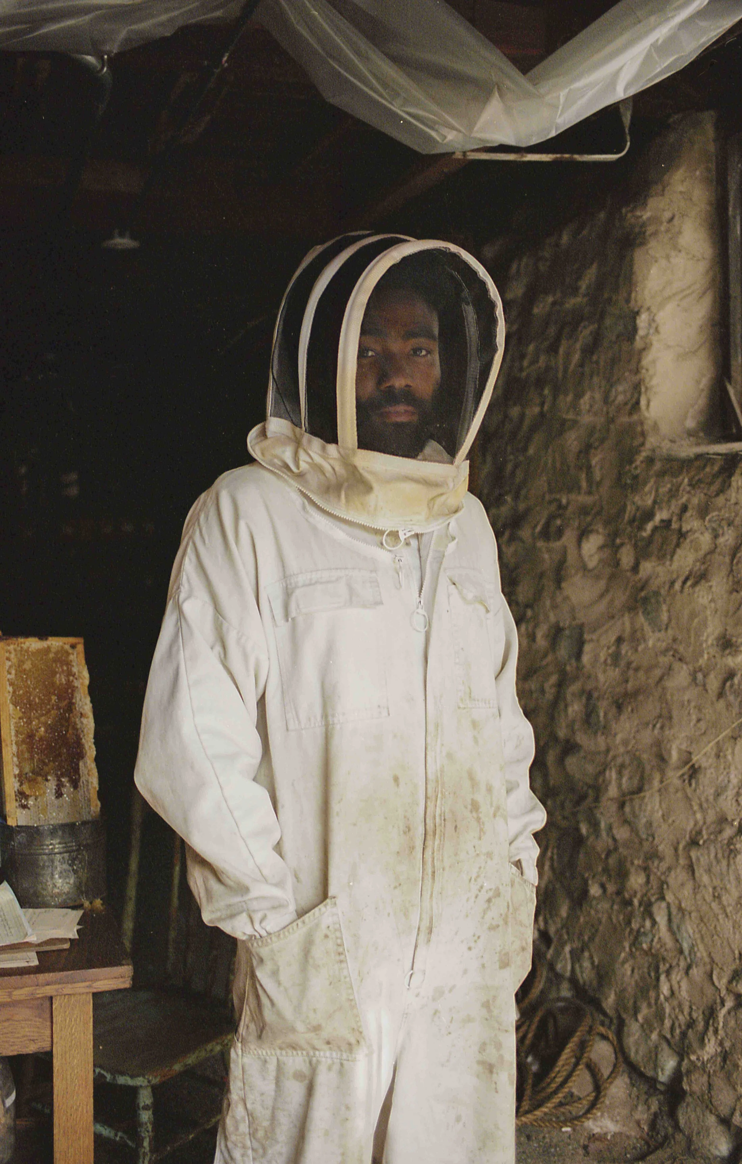 A man wearing a white beekeeping suit and a mesh veil over his face stands indoors near a stone wall, with some ropes and a wooden table in the background.