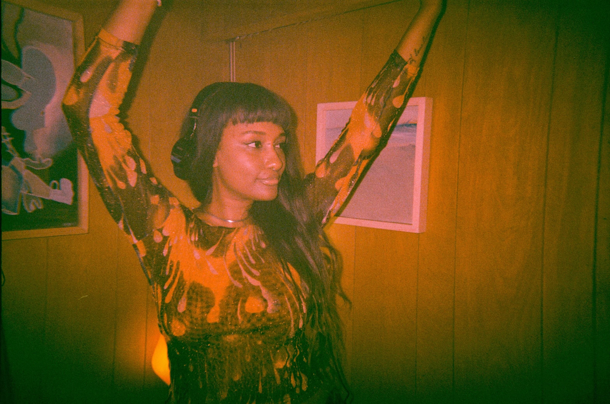 A woman with short hair is wearing headphones and a long-sleeved dress with an orange and black pattern, posing with her arms raised against a wooden wall with colorful framed pictures.