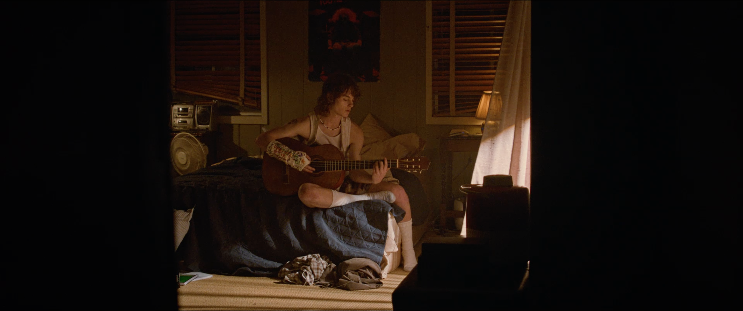 A young person with curly hair sitting on a bed, playing an acoustic guitar in a dimly lit bedroom, surrounded by posters, a small stereo, and a fan.