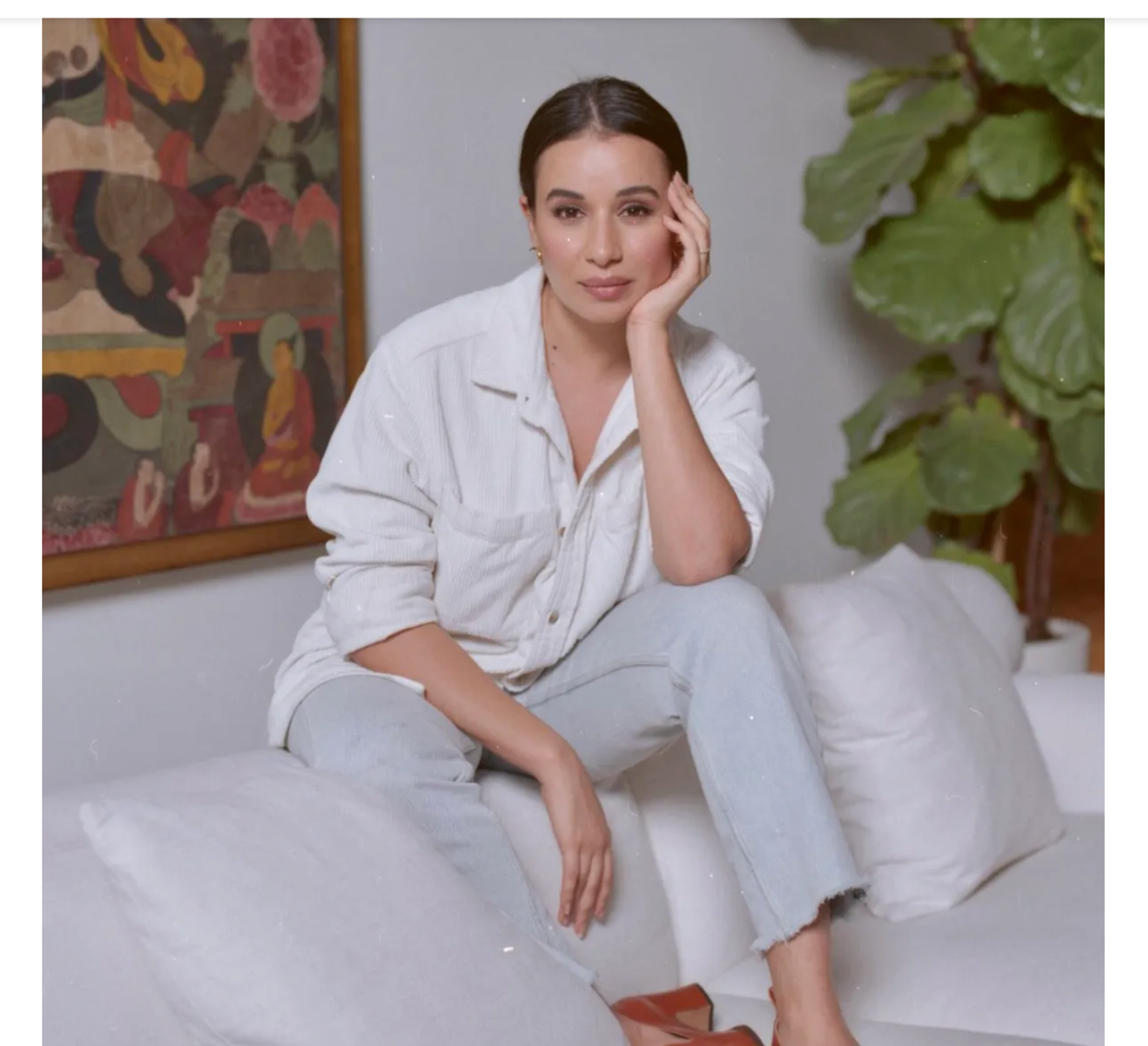 Sylvia Zakhary, A woman with dark hair sitting on a white sofa, resting her face on her hand, wearing a white shirt and light jeans, in a room decorated with a large plant, a framed artwork, and white cushions.