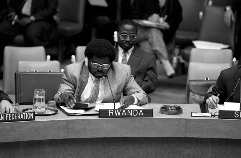 A man with glasses and an afro hairstyle sitting at a conference table labeled 'RWANDA,' reading papers and using a tablet. Other people are seated in the background.
