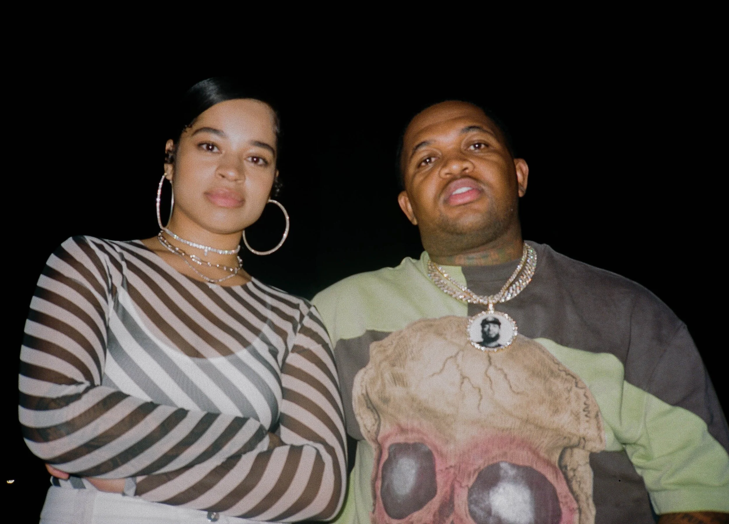 A woman with long black hair, wearing hoop earrings, layered necklaces, and a black and white striped top, standing next to a man with short hair, wearing jewelry and a T-shirt featuring a face illustration against a dark background.