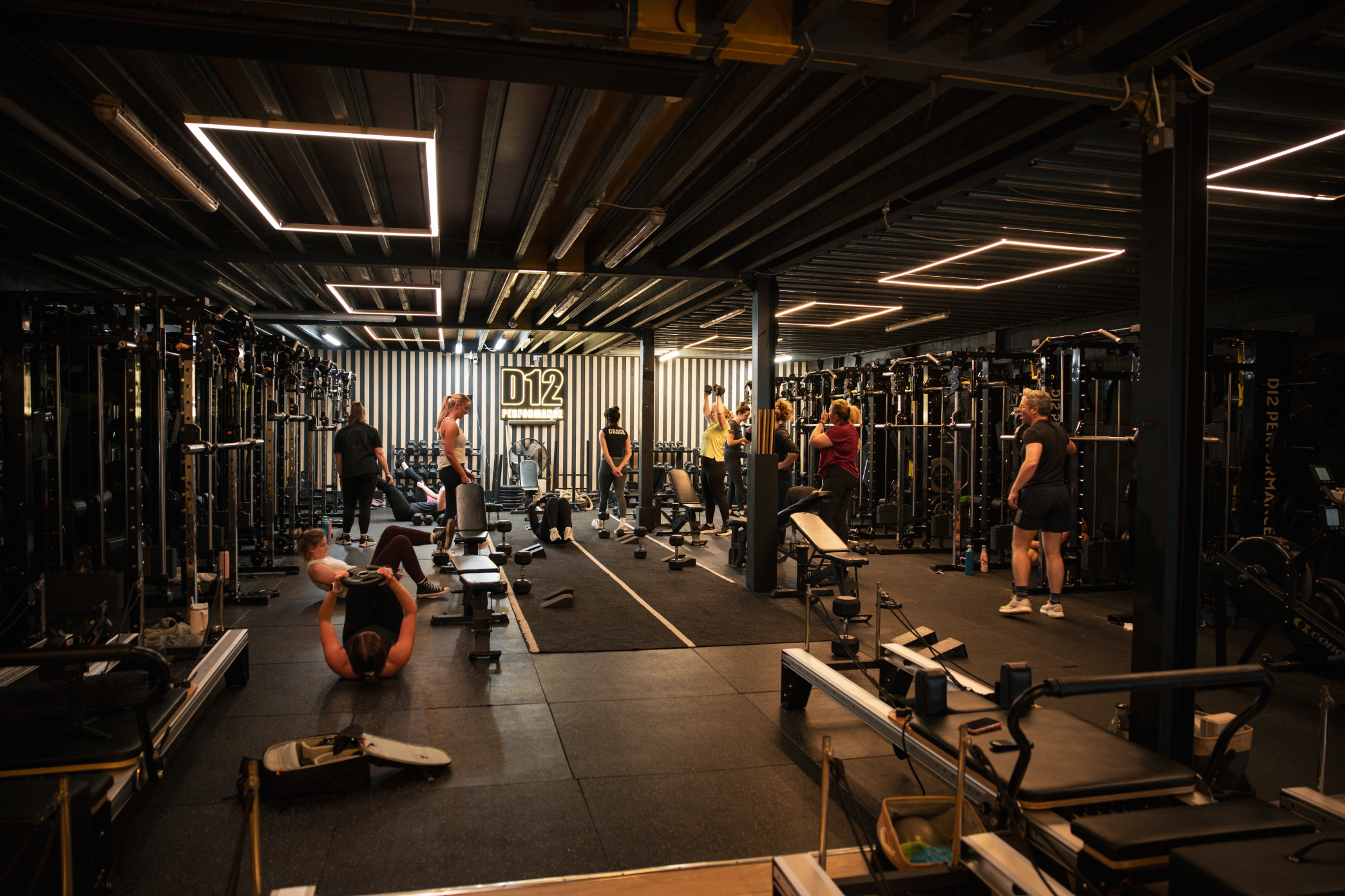 A busy gym with people working out using various equipment, including weights and benches, with a black and white striped wall and a sign reading 'D12 Performance' in the background.