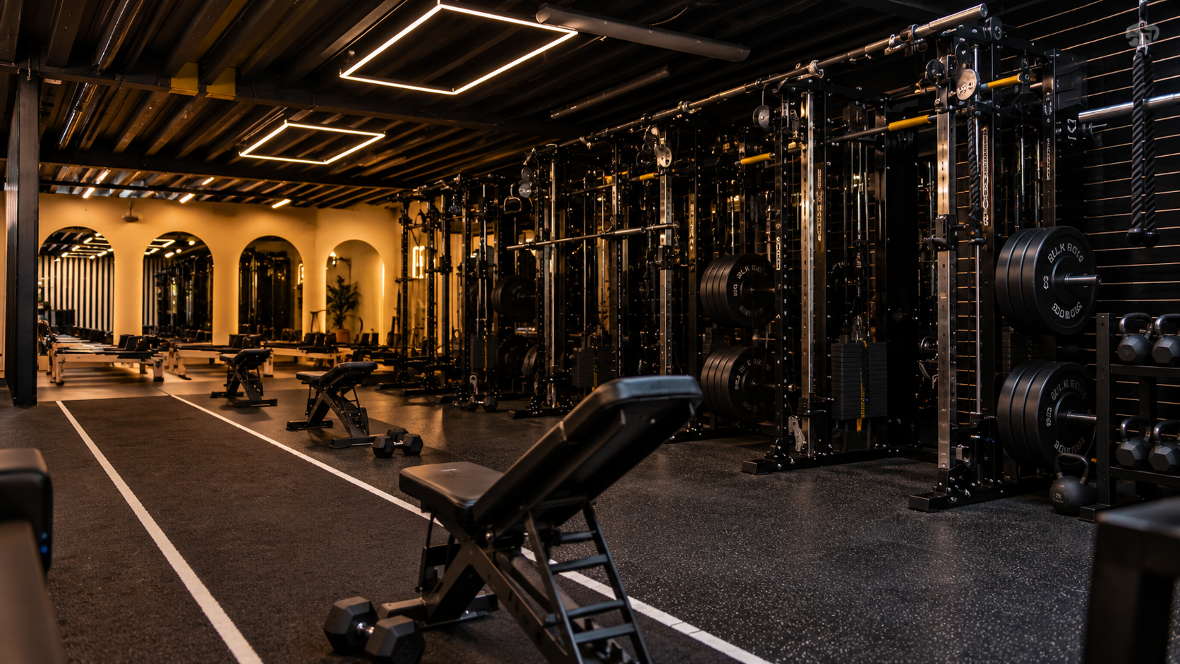 A modern gym with black equipment, weight racks, benches, dumbbells, and kettlebells, with a black ceiling and warm lighting.