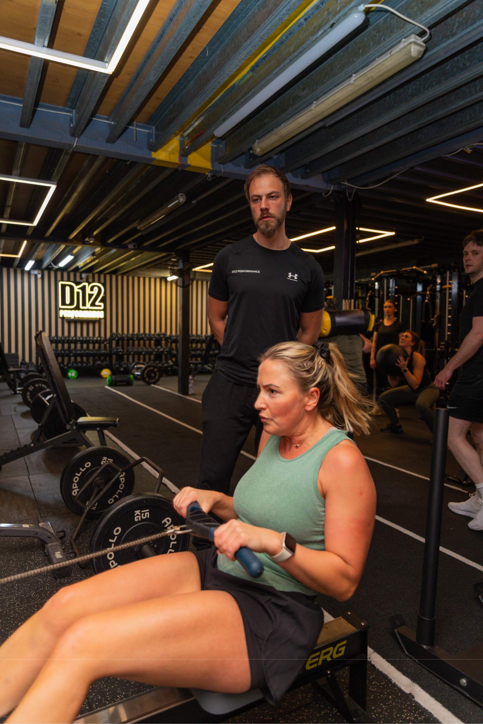 Multiple people workout in a modern gym, featuring a woman using gym equipment, a man standing behind her, and others in the background exercising. The gym has black and yellow decor, with a sign reading 'D12 PERFORMANCE'.