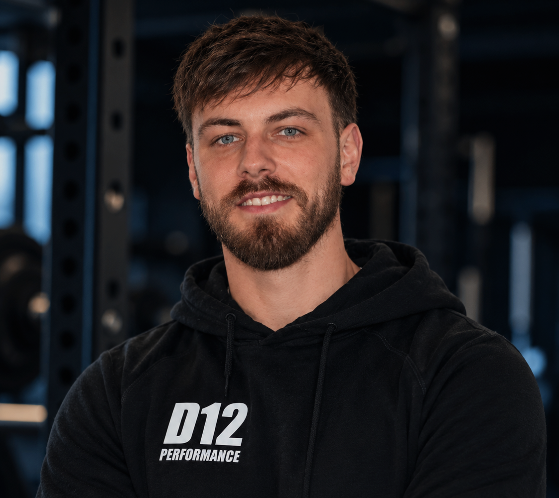 A young man with brown hair and a beard, wearing a black hoodie with 'D12 PERFORMANCE' written on it, smiling in a gym setting with weightlifting equipment in the background.