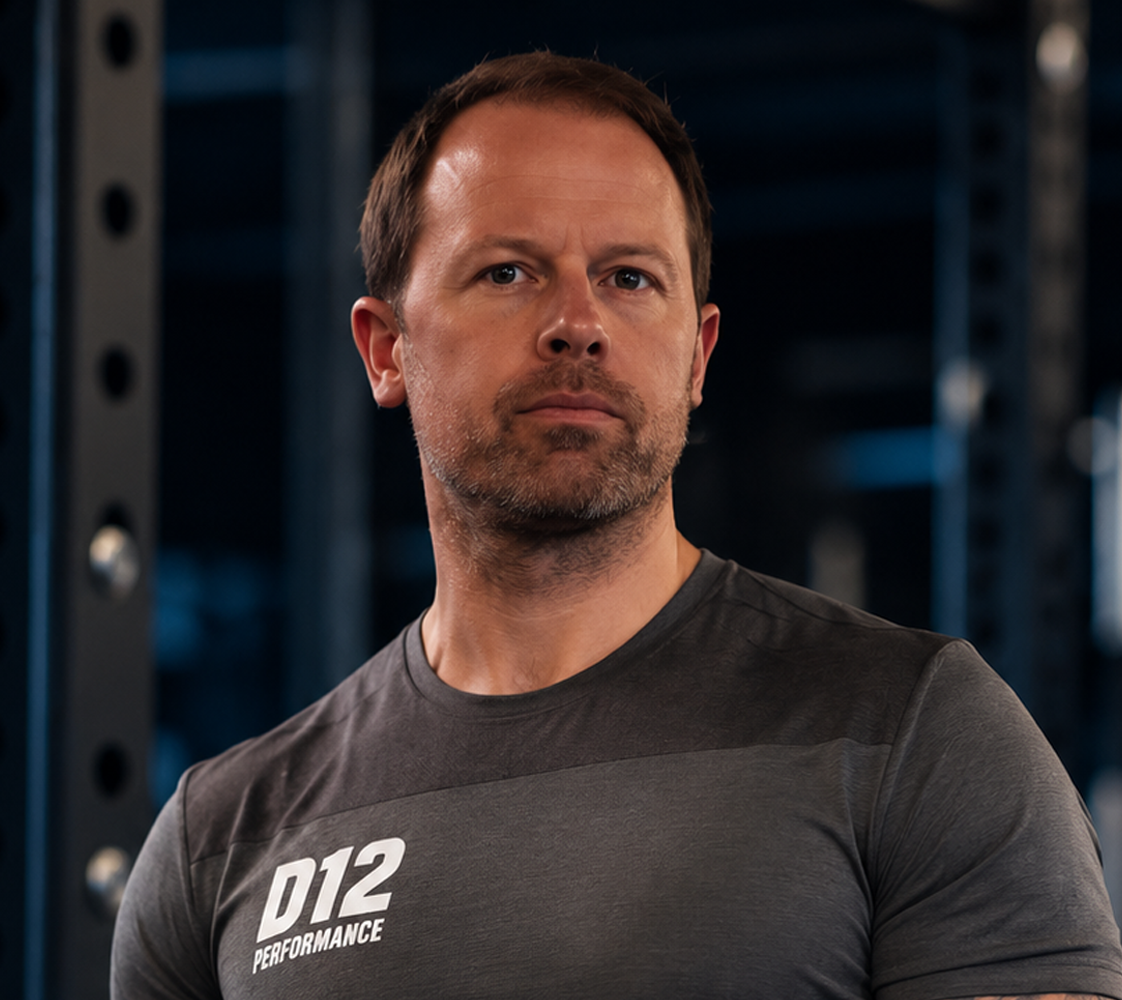 A man standing in a gym, wearing a black athletic shirt with 'D12 PERFORMANCE' printed on it, looking serious, with gym equipment in the background.