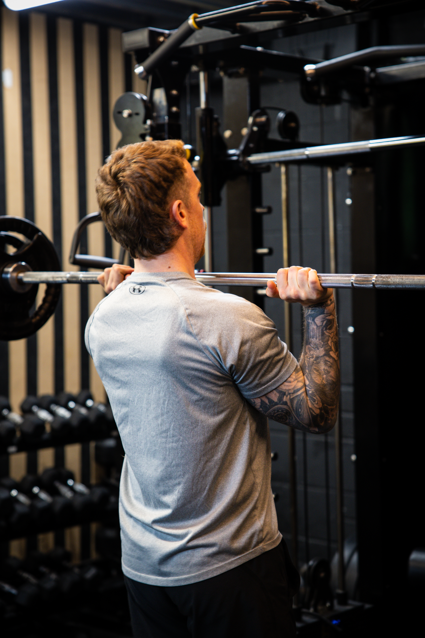 A man with tattoos on his right arm lifting a barbell during a workout in a gym.