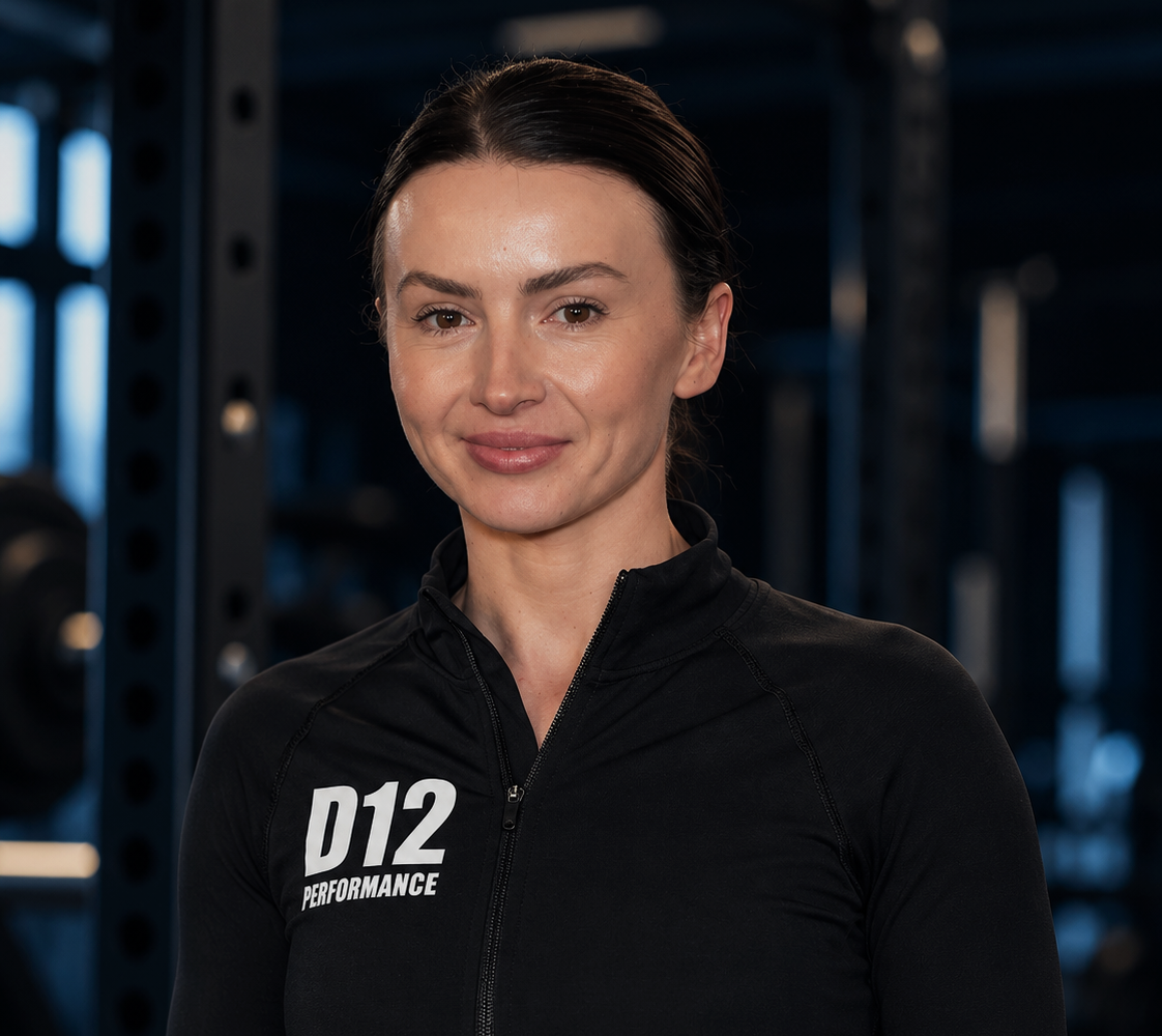 Close-up of a woman in a black athletic jacket with 'D12 PERFORMANCE' logo, smiling confidently in a gym setting with weight racks in the background.
