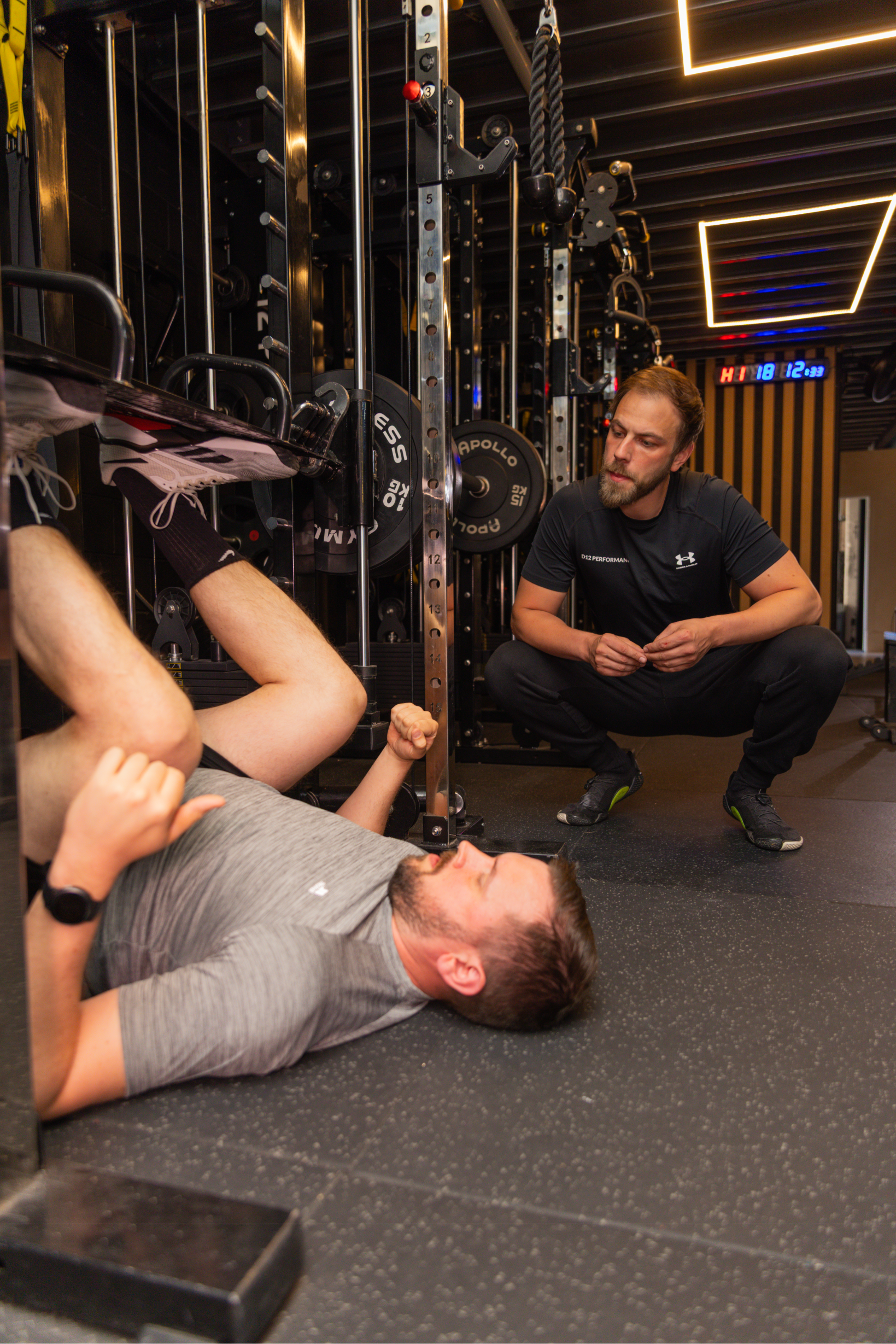 A man lying on the gym floor during a workout, with a trainer crouching nearby, in a fitness center with weightlifting equipment and a digital clock on the wall.