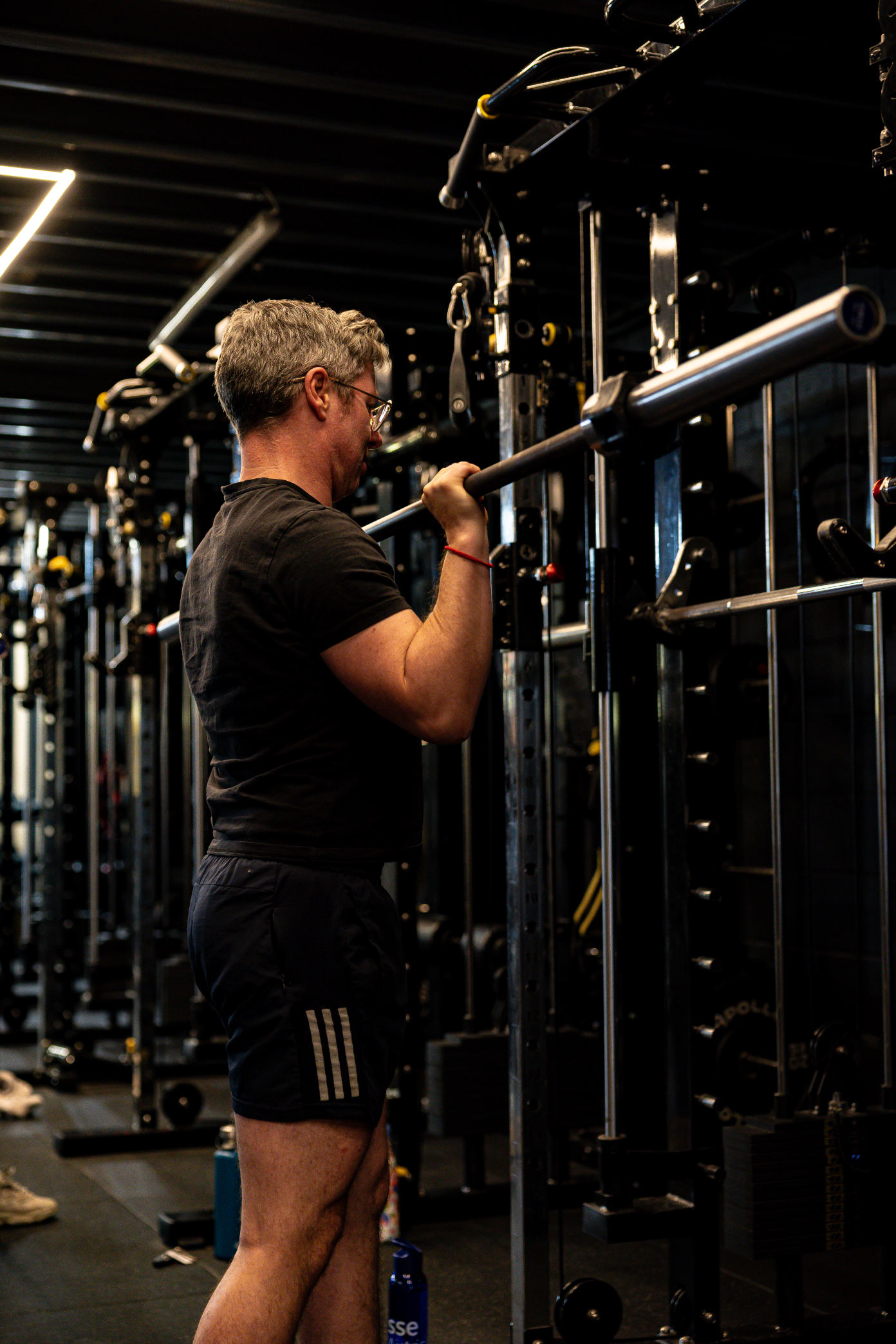 A man with glasses, wearing a black t-shirt and black shorts, performing a barbell squat in a gym, standing in front of a squat rack surrounded by various gym equipment.
