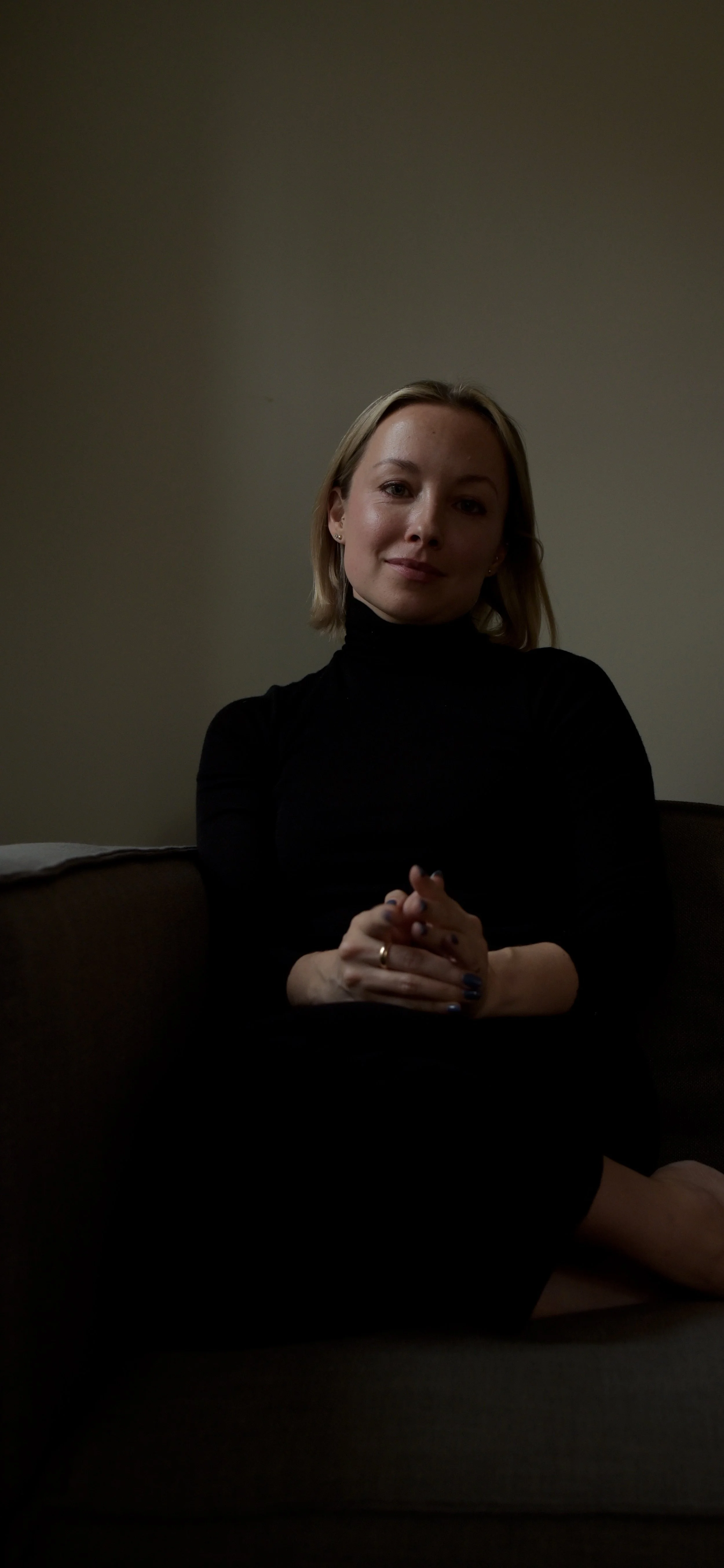 A woman with blonde hair, wearing a black turtleneck, sitting on a brown couch against a plain wall, with her hands clasped together in front of her.