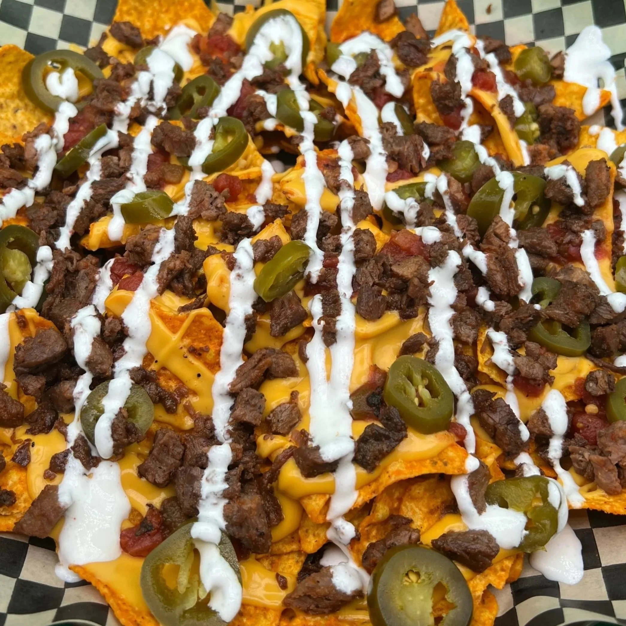A close-up of two loaded nachos, topped with chopped beef, jalapeño slices, sour cream, and melted cheese on a checkered black and white tray.