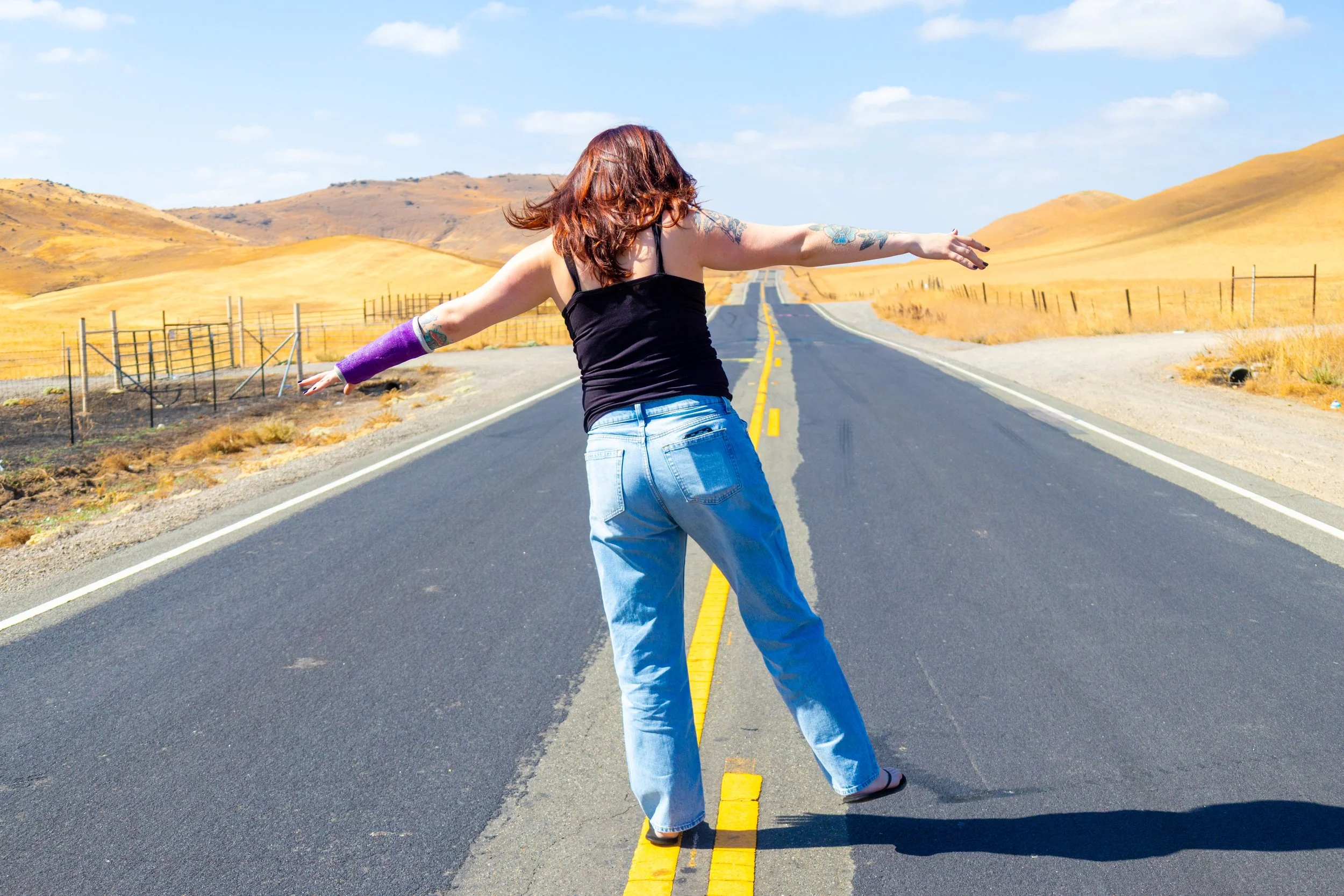 A person with red hair and tattoos on their arms is standing in the middle of an empty two-lane road with yellow markings, stretching their arms wide open. They are wearing a black tank top, blue jeans, and sandals. The landscape features rolling hil