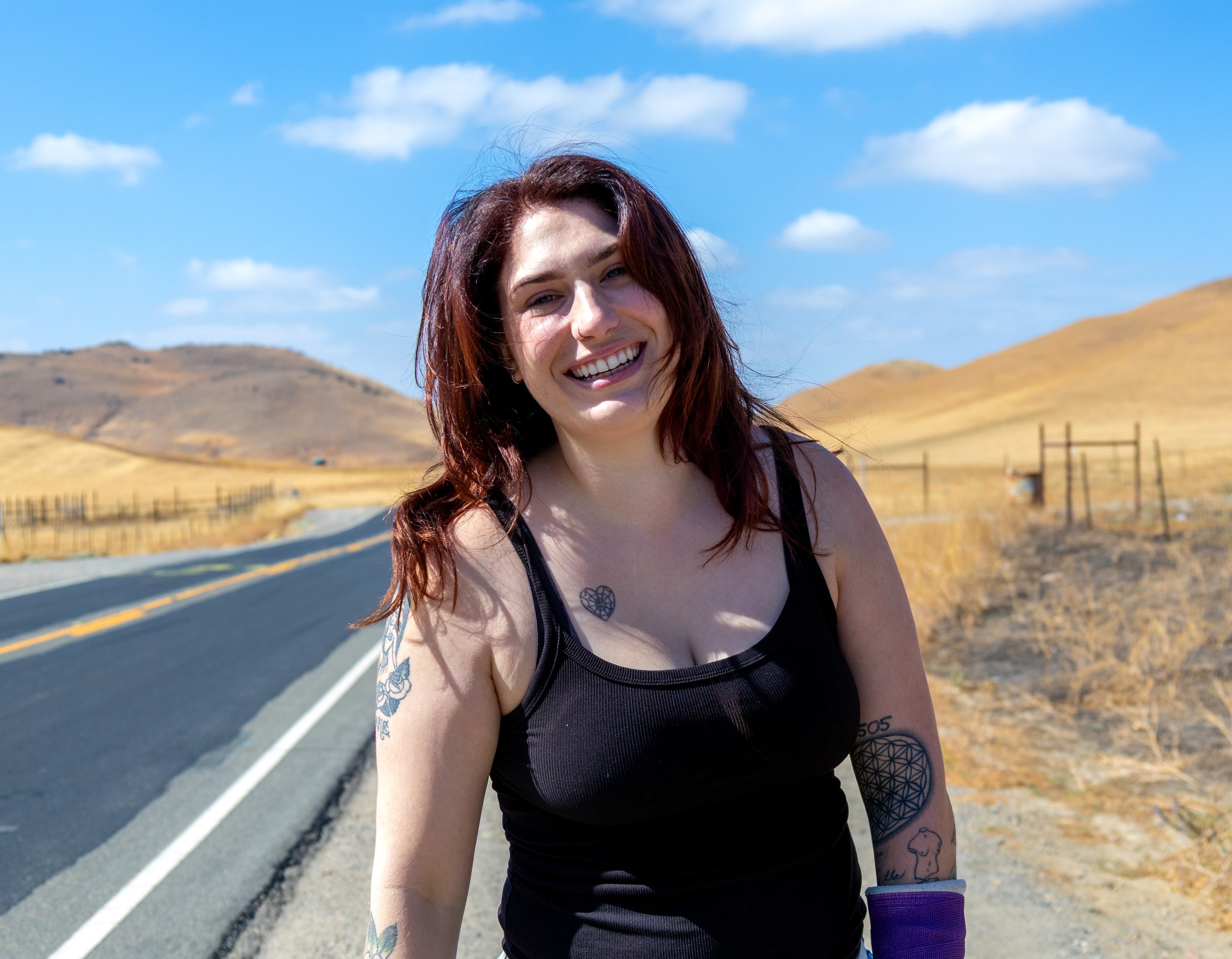 A smiling woman with tattoos on her arm and shoulder stands outdoors on a sunny day near a rural road with hills and a wooden fence in the background.