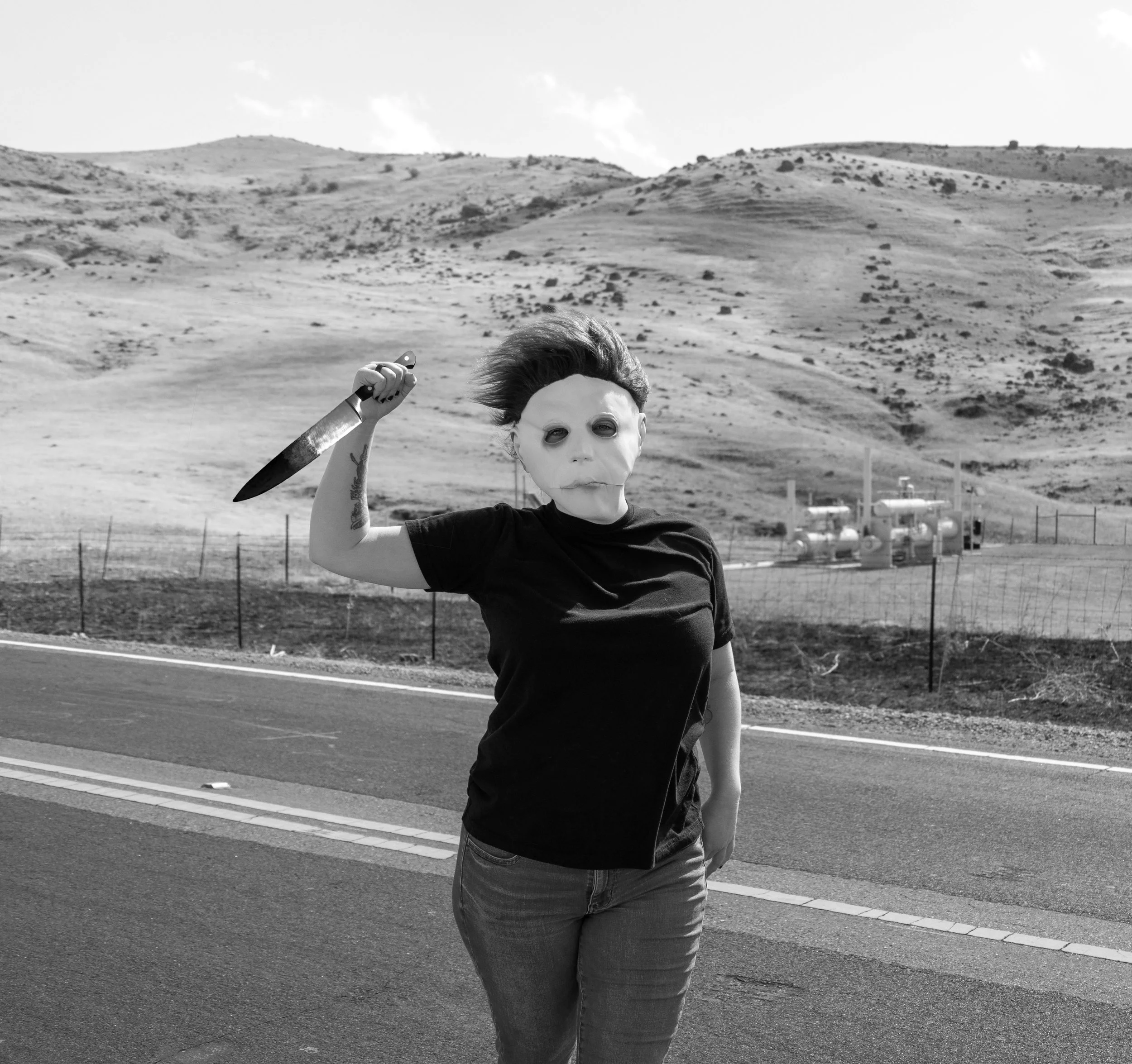 A person with a painted face, wearing a black shirt and jeans, holding a rebellious posture, standing on the side of a road in an arid landscape with hills in the background. The person has short hair and tattoos on their arm.