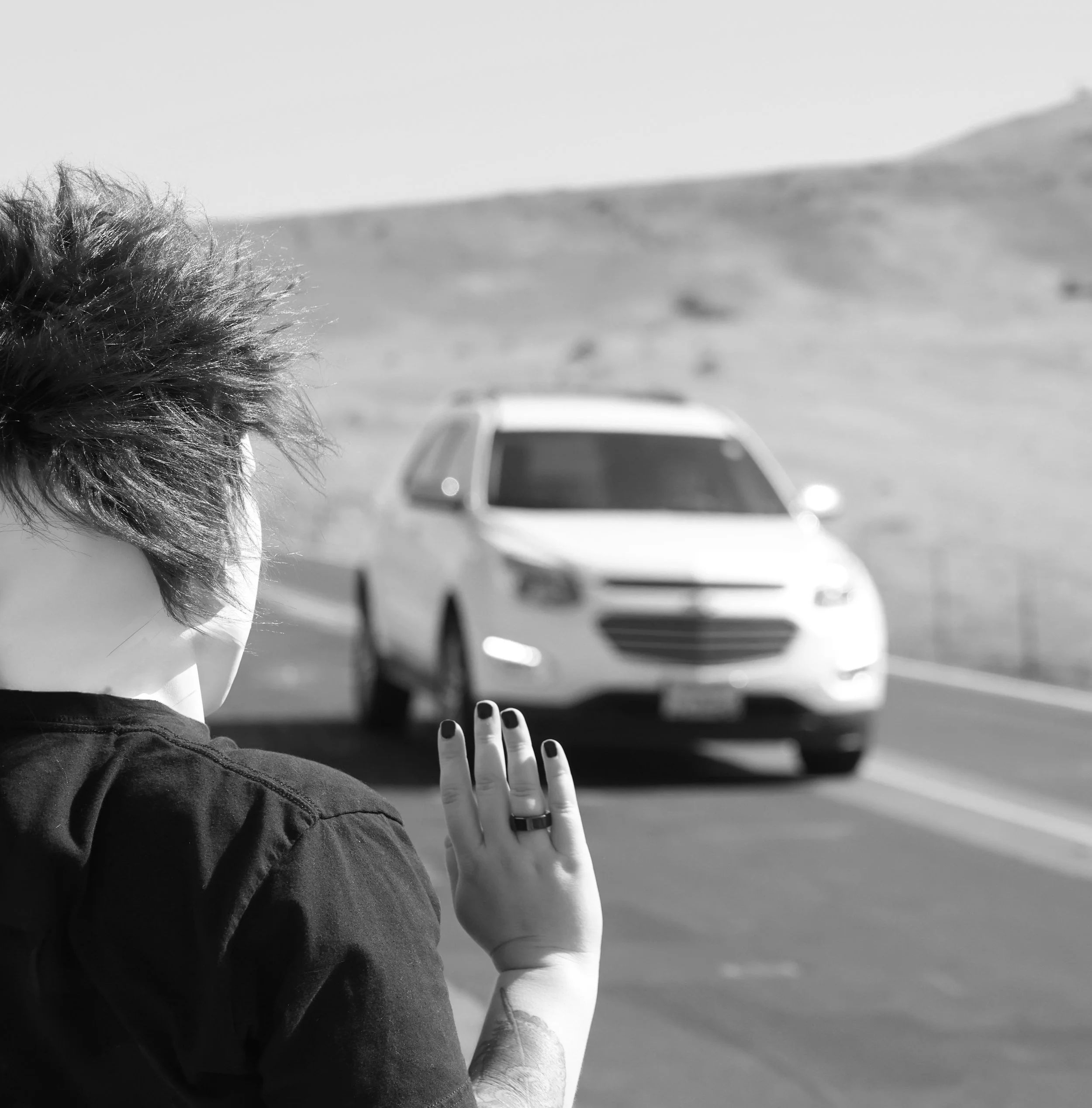 Black and white photo of a person with short hair and painted nails, touching a window with a blurred car in the background.