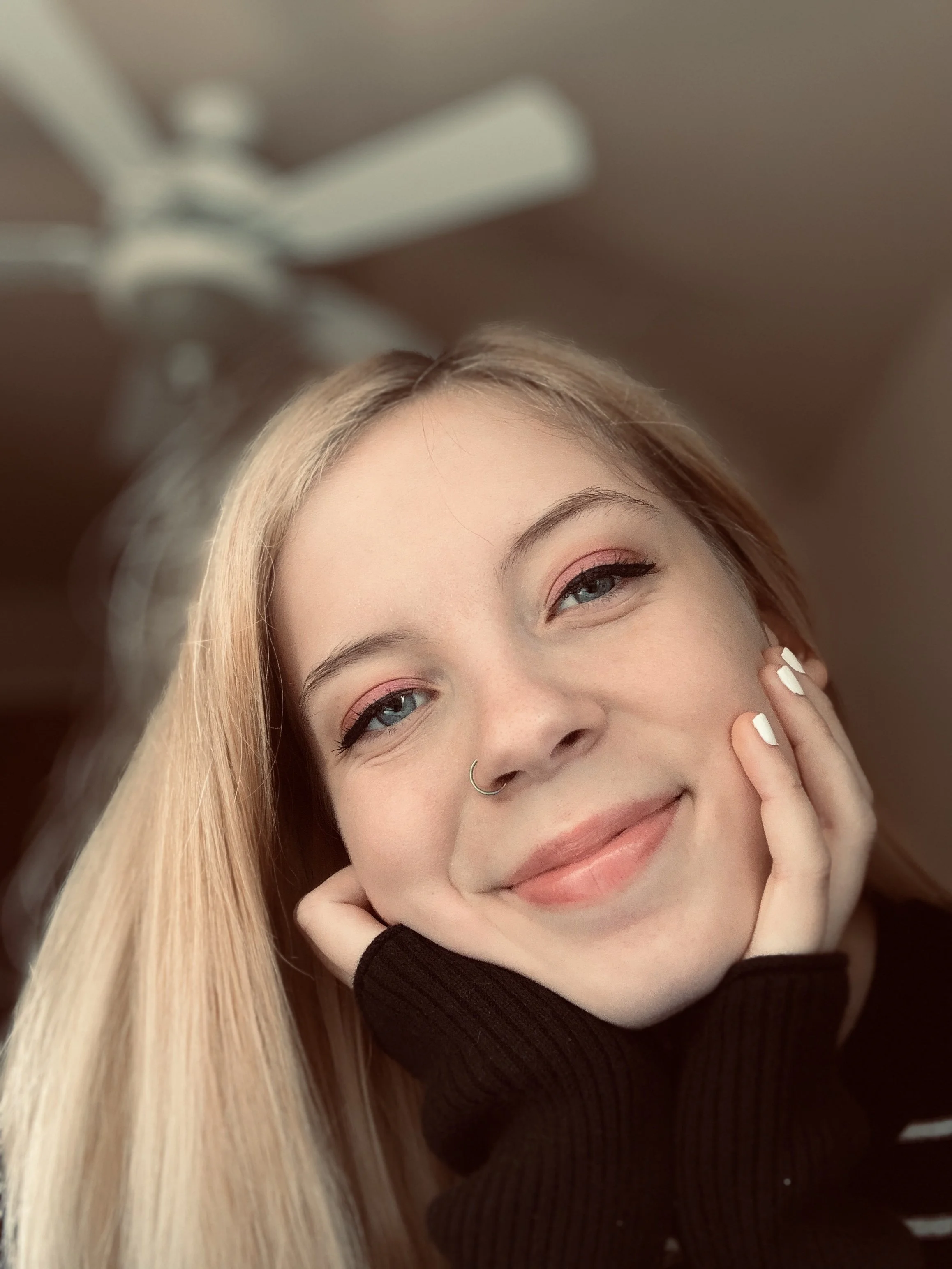 Close-up photo of a young woman with blonde hair and blue eyes, smiling gently, with her hand resting on her cheek and wearing a black top.