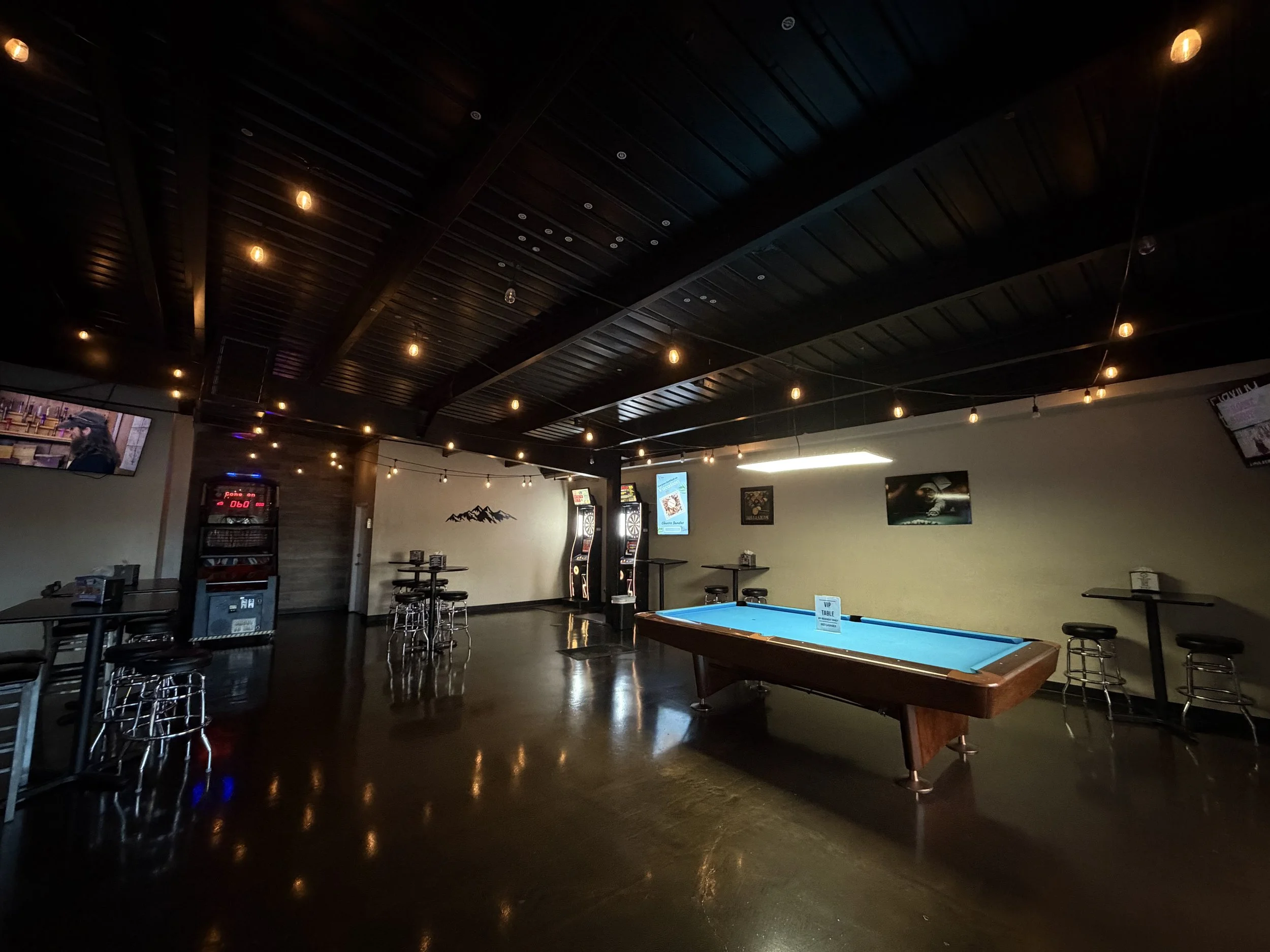 A dimly lit game room with a pool table in the center, dark wood ceiling with hanging string lights, and electronic dartboards on the wall.
