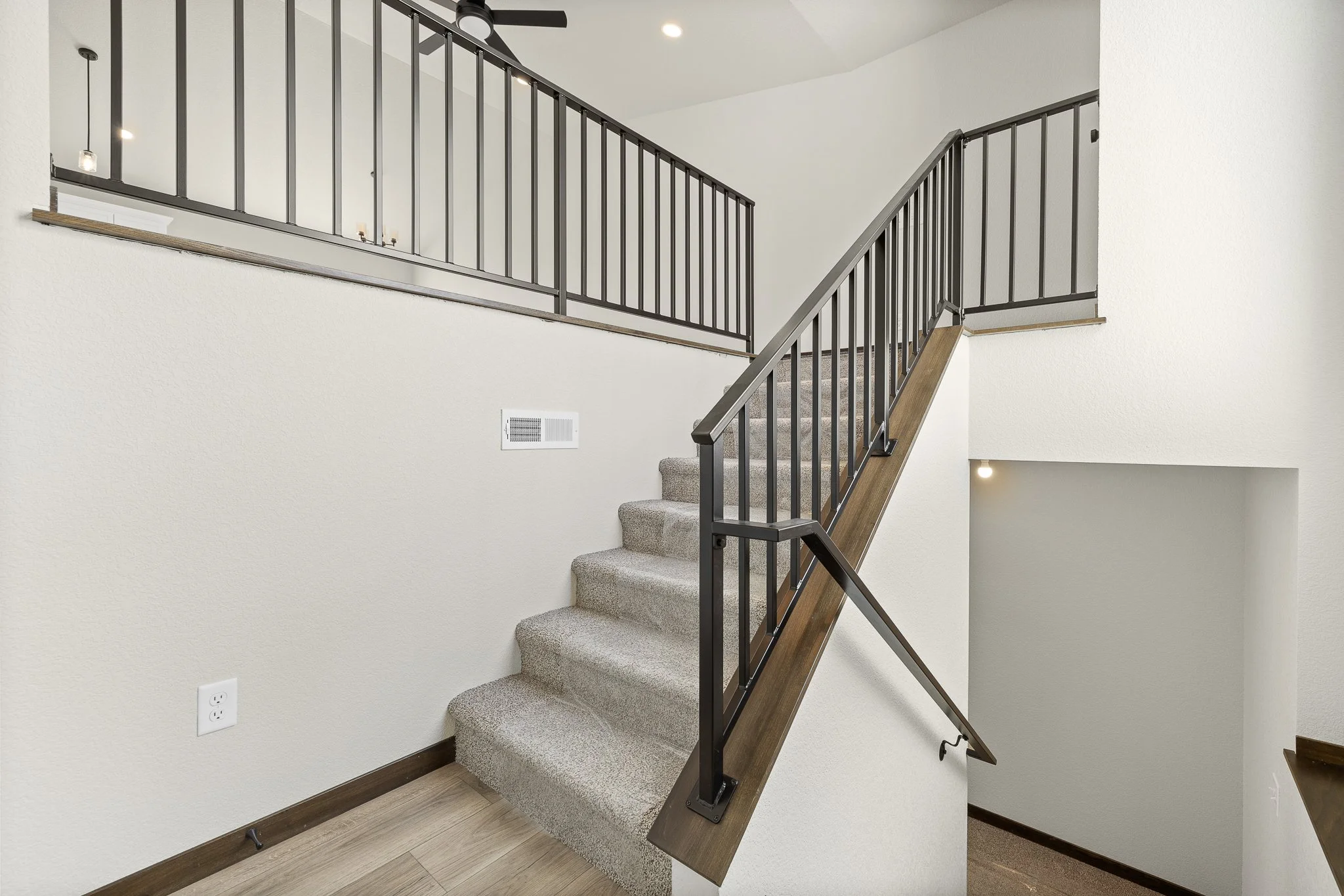 Interior view of a staircase with beige carpeted steps, black metal railing, and wooden handrail. White textured walls, ceiling with recessed lighting, ceiling fan, and an electrical outlet visible.